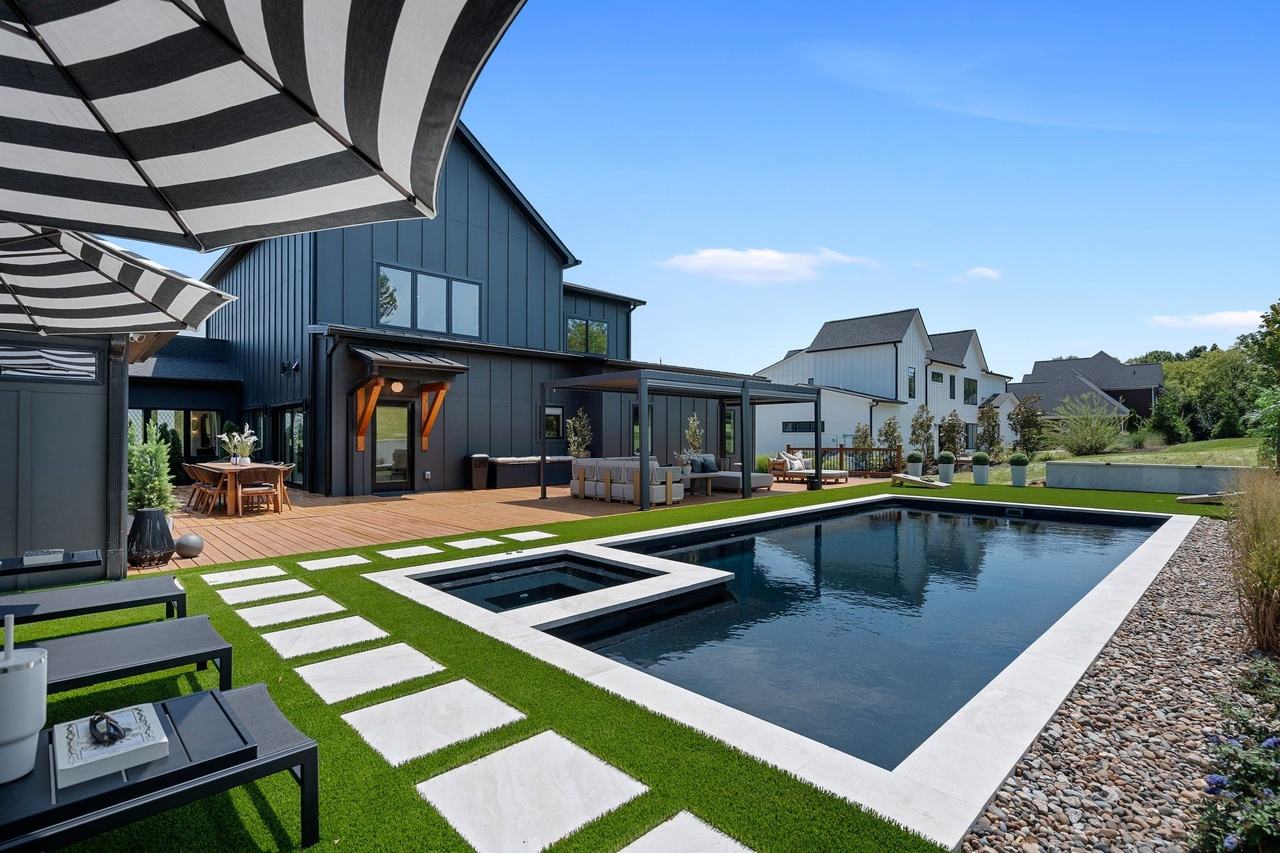The pool from the patio toward the house: a dark pool with a built-in spa and travertine coping, pavers set in turf, and the louvered pergola over the deck.