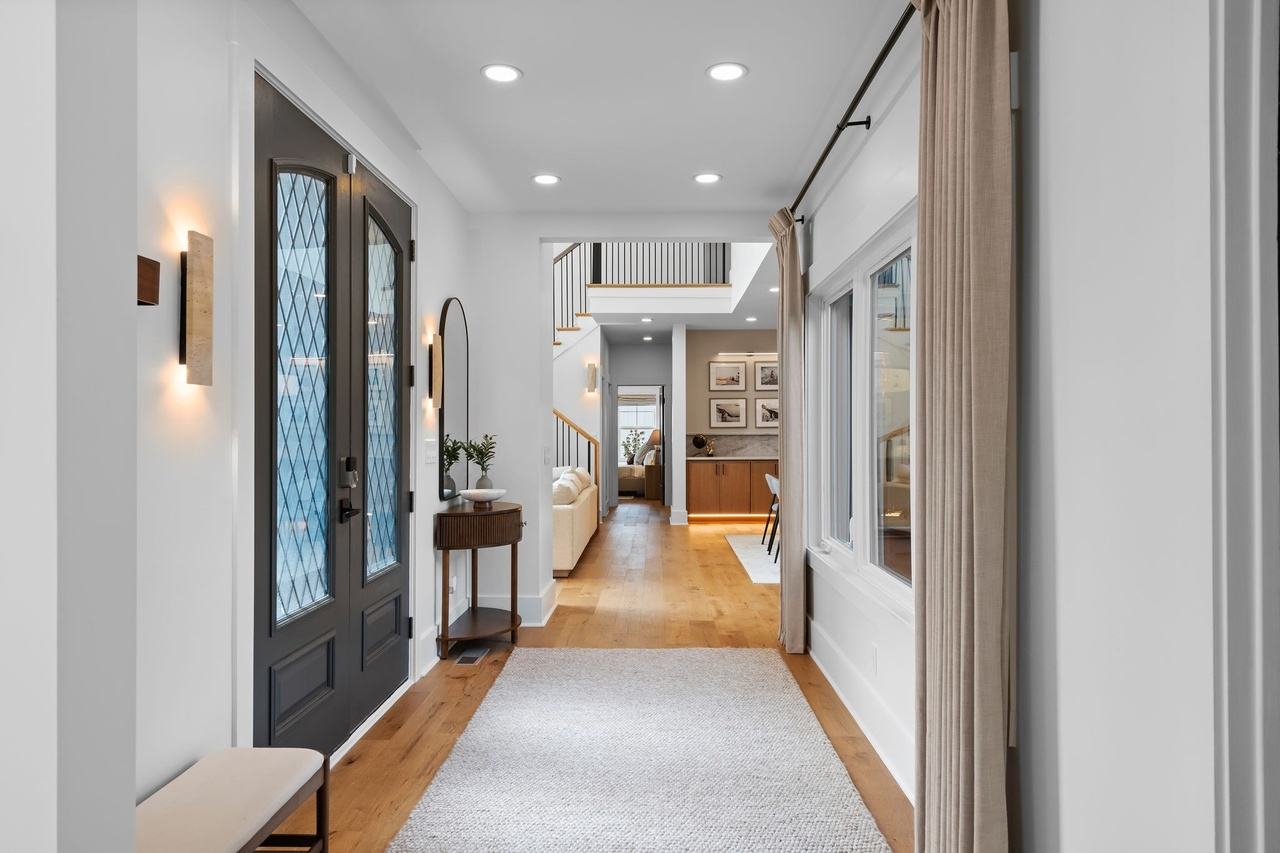 The foyer looking down the entry hall: dark arched double doors with leaded glass, travertine sconces, a woven runner and linen curtains on a black rod.