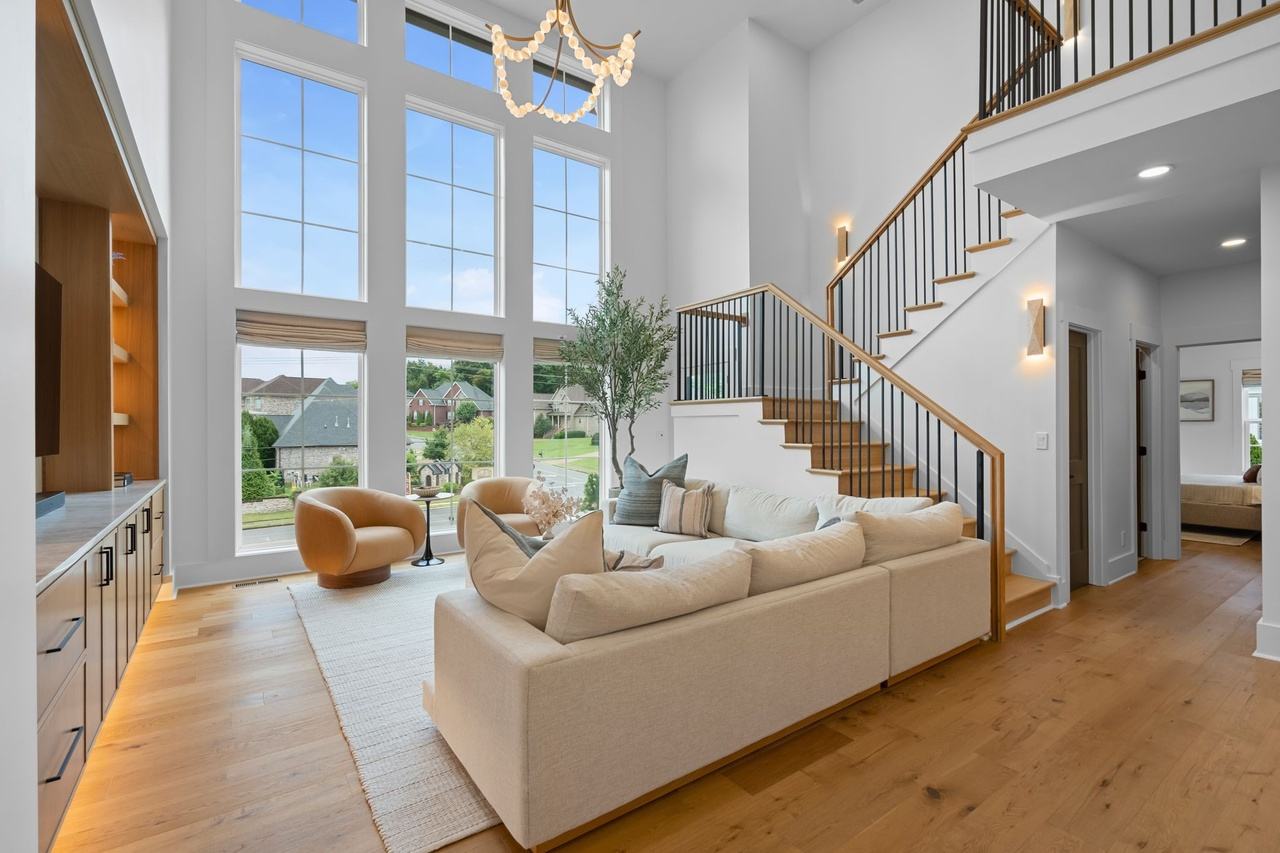 The two-story living room: a cream sectional facing the two-story wall of windows, an oak stair with black balusters, and a beaded garland chandelier overhead.