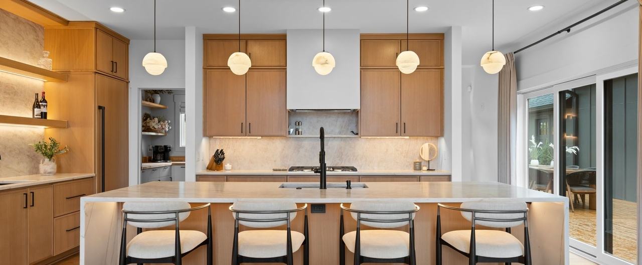 The kitchen island head on: a quartz waterfall top, four black-framed counter stools, five alabaster globe pendants and oak cabinetry either side of a white hood.