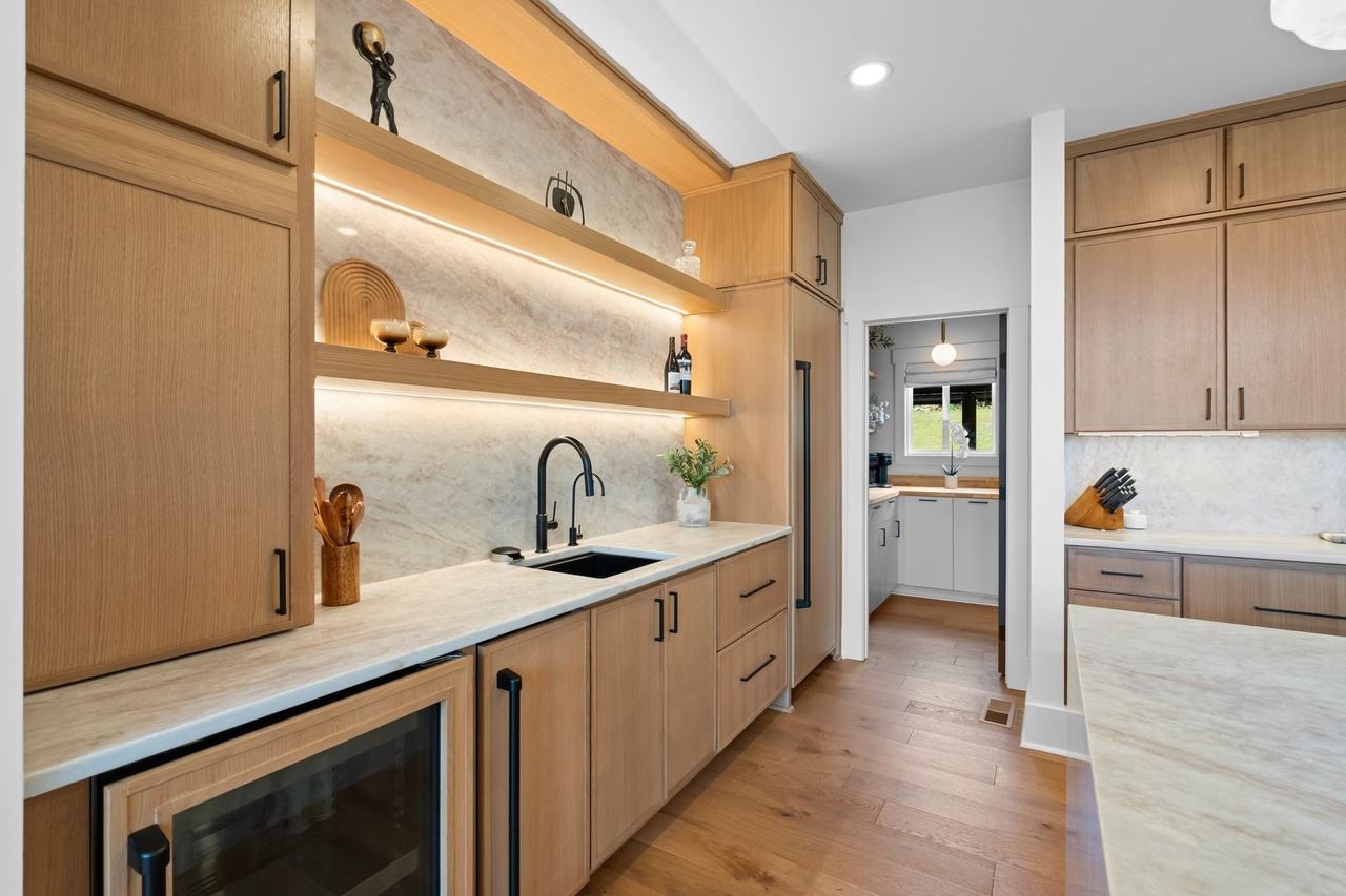 The wet bar: oak cabinets, a glass-front beverage fridge and a full-height quartz splash behind two lit floating shelves.