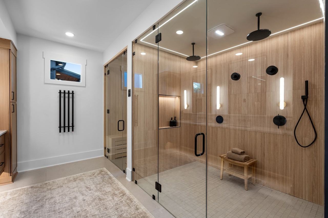 The primary shower: a glass enclosure wrapped in ribbed wood-look tile, two black rain heads, vertical sconces, a lit niche and a teak bench, with the sauna door at left.