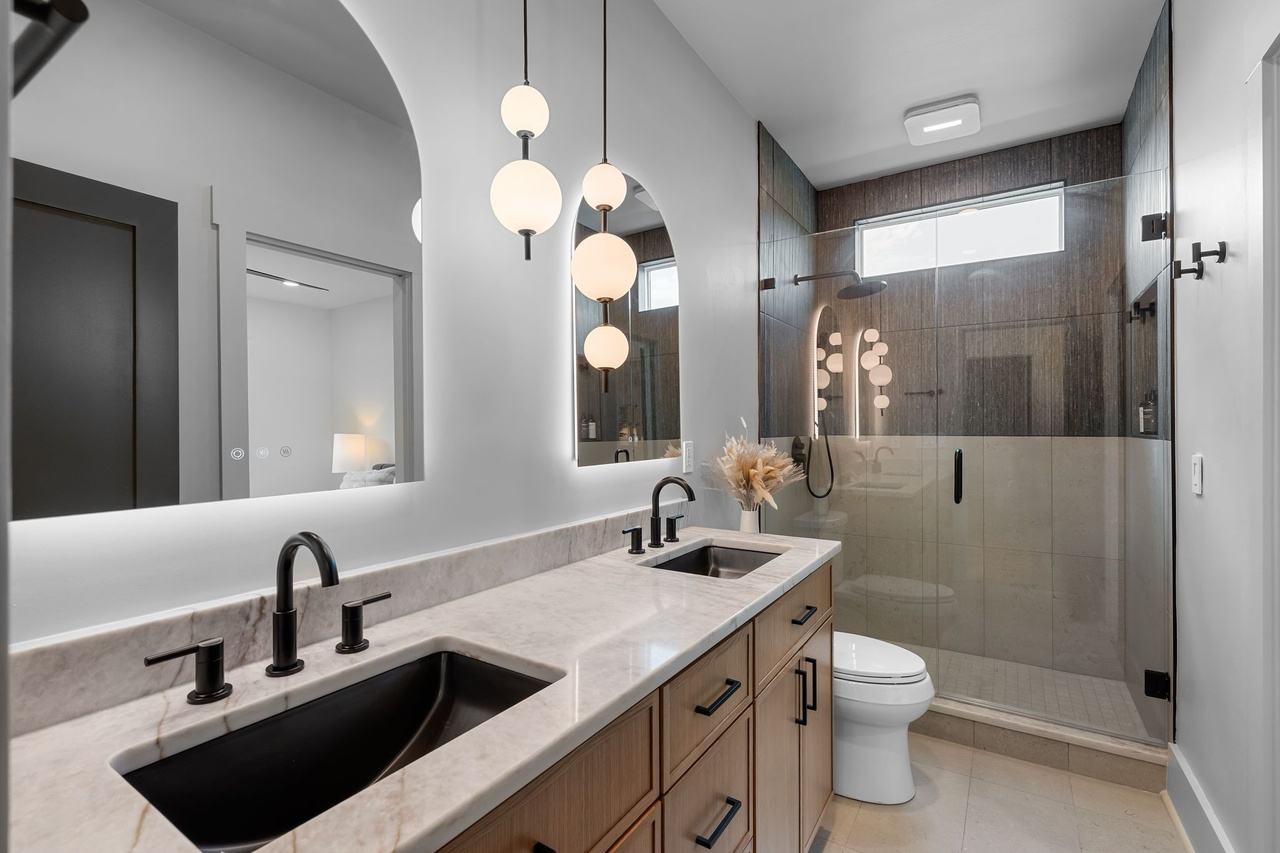 The upstairs bath: a double vanity with black faucets, arched backlit mirrors, globe pendants and a brown tile shower.