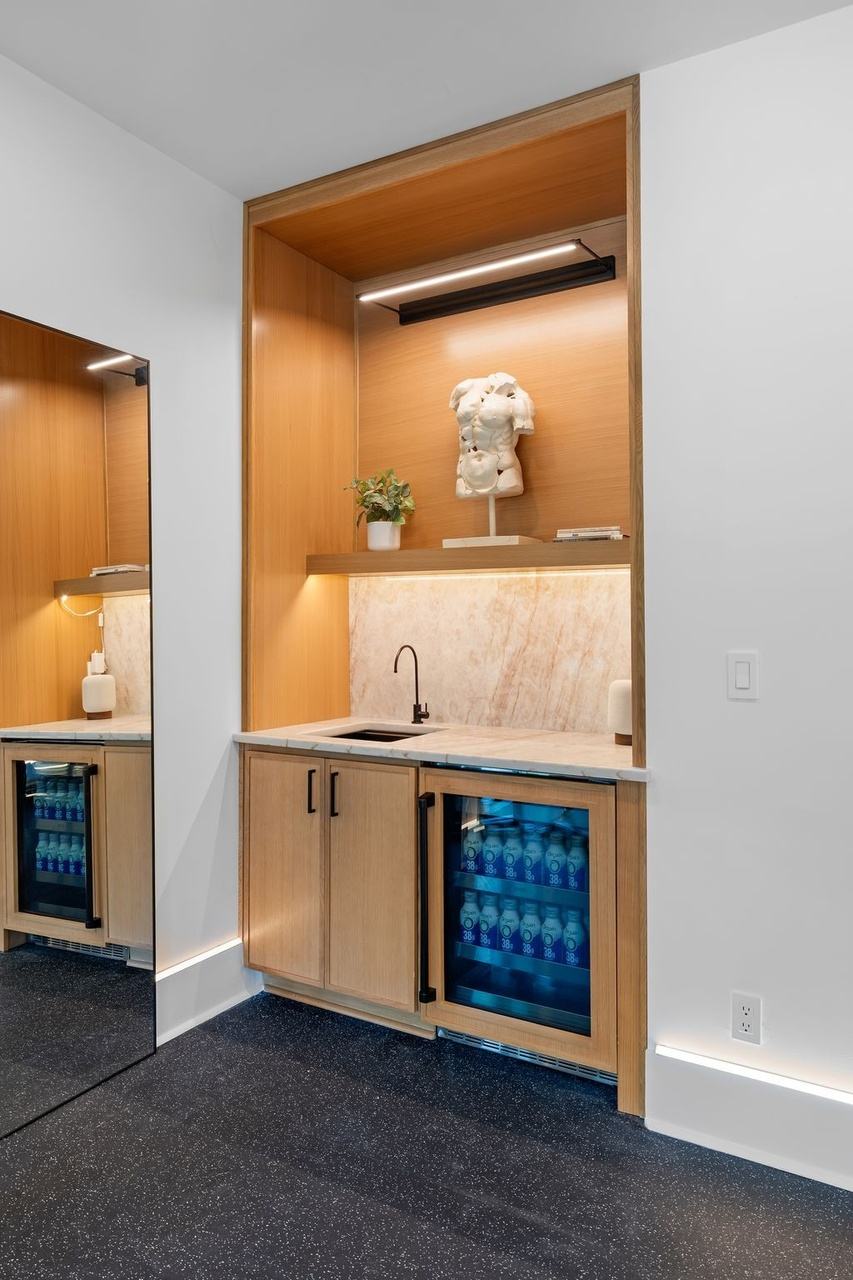 The gym wet bar: an oak niche with a small sink, a stone splash and a glass-front beverage refrigerator.