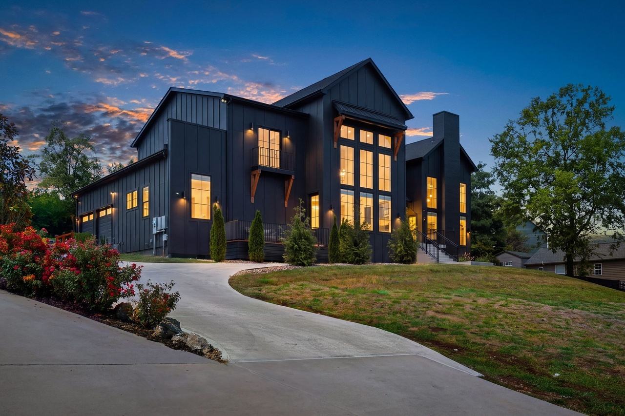 The house at dusk from the curving driveway: black board-and-batten siding, a two-story wall of windows lit from within, cedar brackets under the eaves.