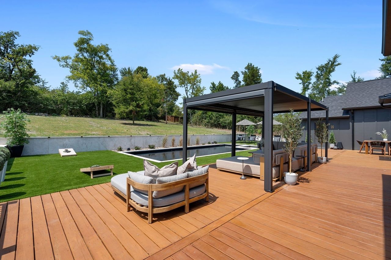 The deck and pergola from the corner of the deck: a curved teak daybed in the foreground, a sectional under the louvered roof, the pool beyond.