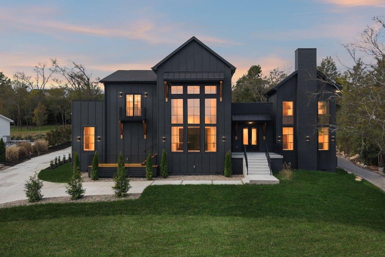 2025 photograph (before): Exterior Front, straight on at dusk, with board and batten, lit glass wall of windows, brick chimney, front steps