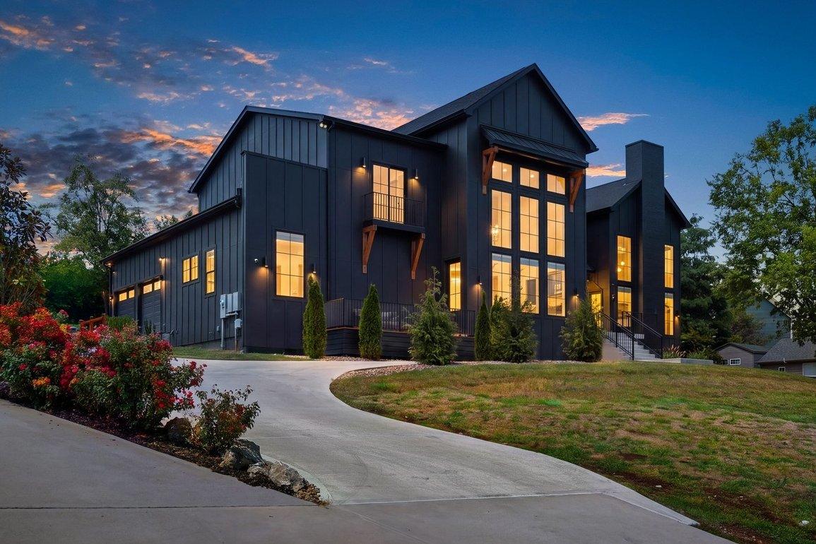 The house at dusk from the curving driveway: black board-and-batten siding, a two-story wall of windows lit from within, cedar brackets under the eaves.