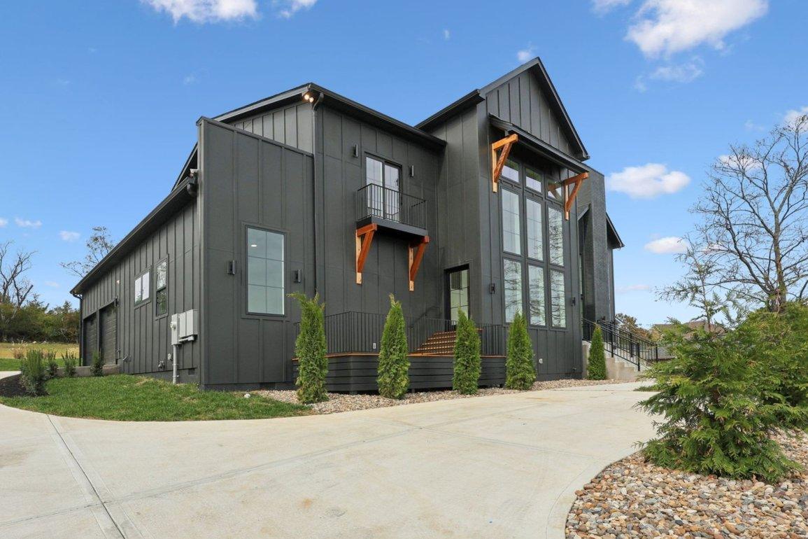 2025 photograph (before): Exterior Front, angled from driveway, garage side, with board and batten, wall of windows, balcony, garage doors