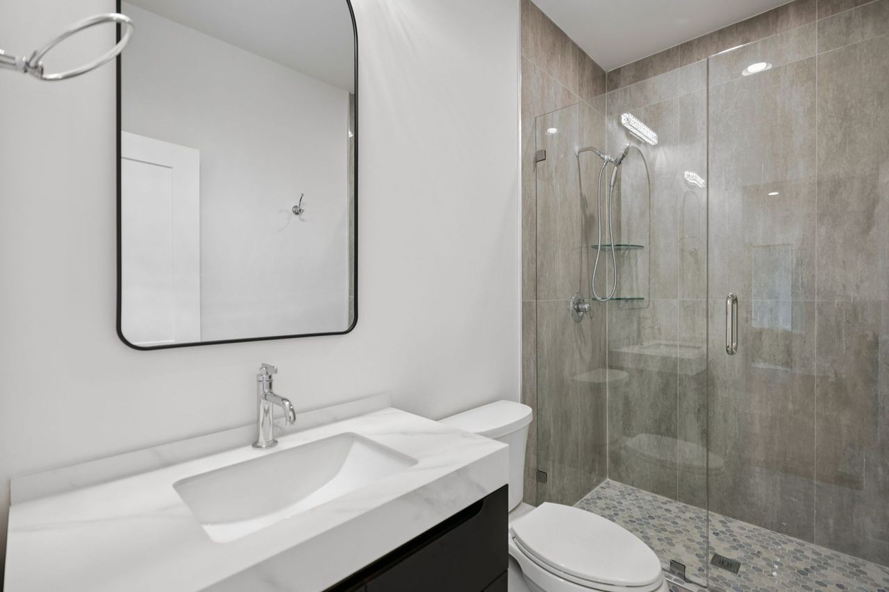 2025 photograph (before): the guest bath, a white vanity top, a black-framed mirror and a gray tile shower.