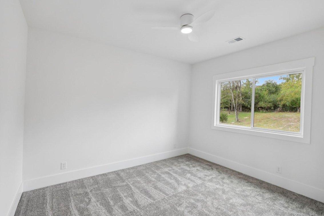 2025 photograph (before): Guest Room 1, toward wide rear window, with wide sliding window, wooded yard view, gray carpet, ceiling fan