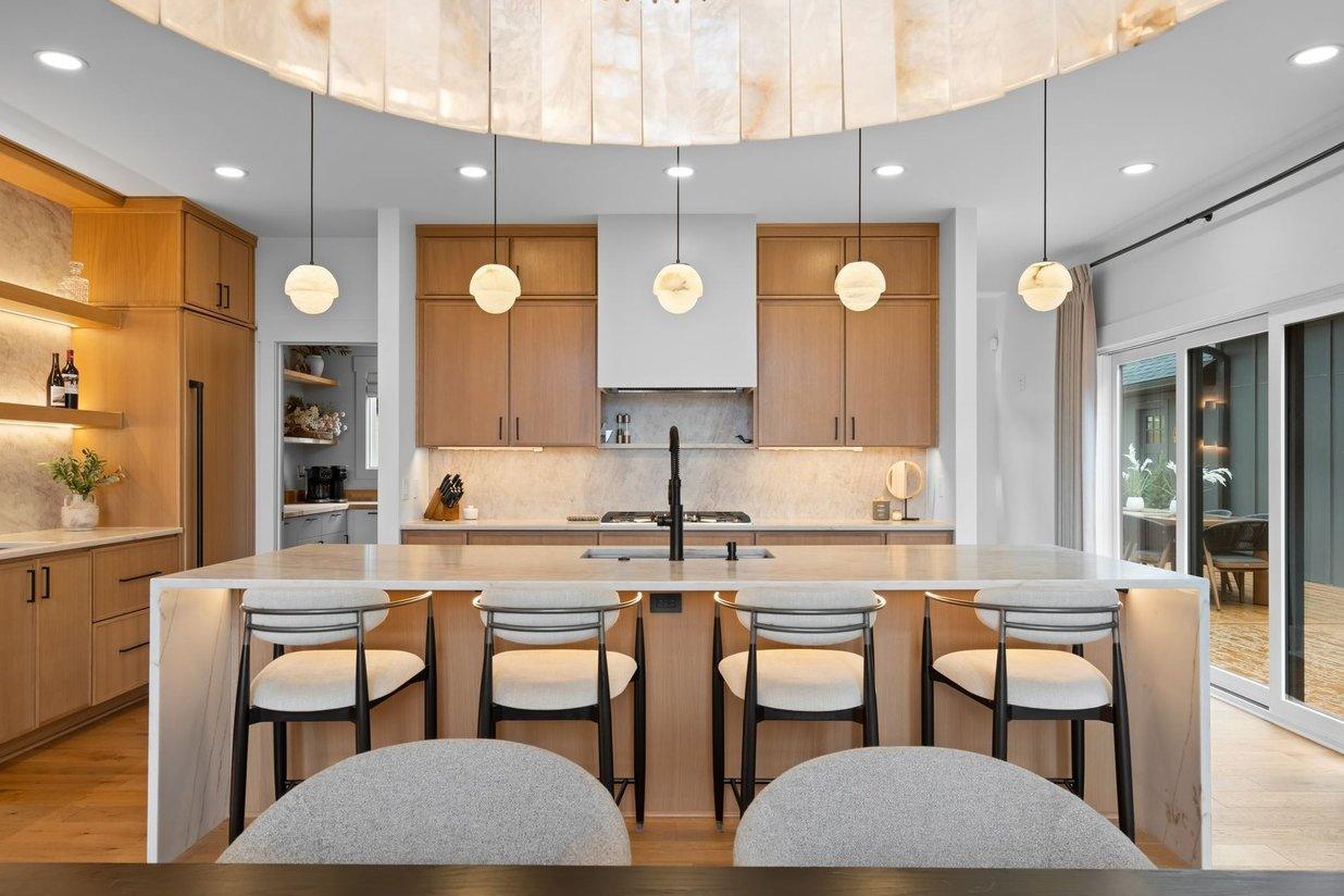 The kitchen island head on: a quartz waterfall top, four black-framed counter stools, five alabaster globe pendants and oak cabinetry either side of a white hood.