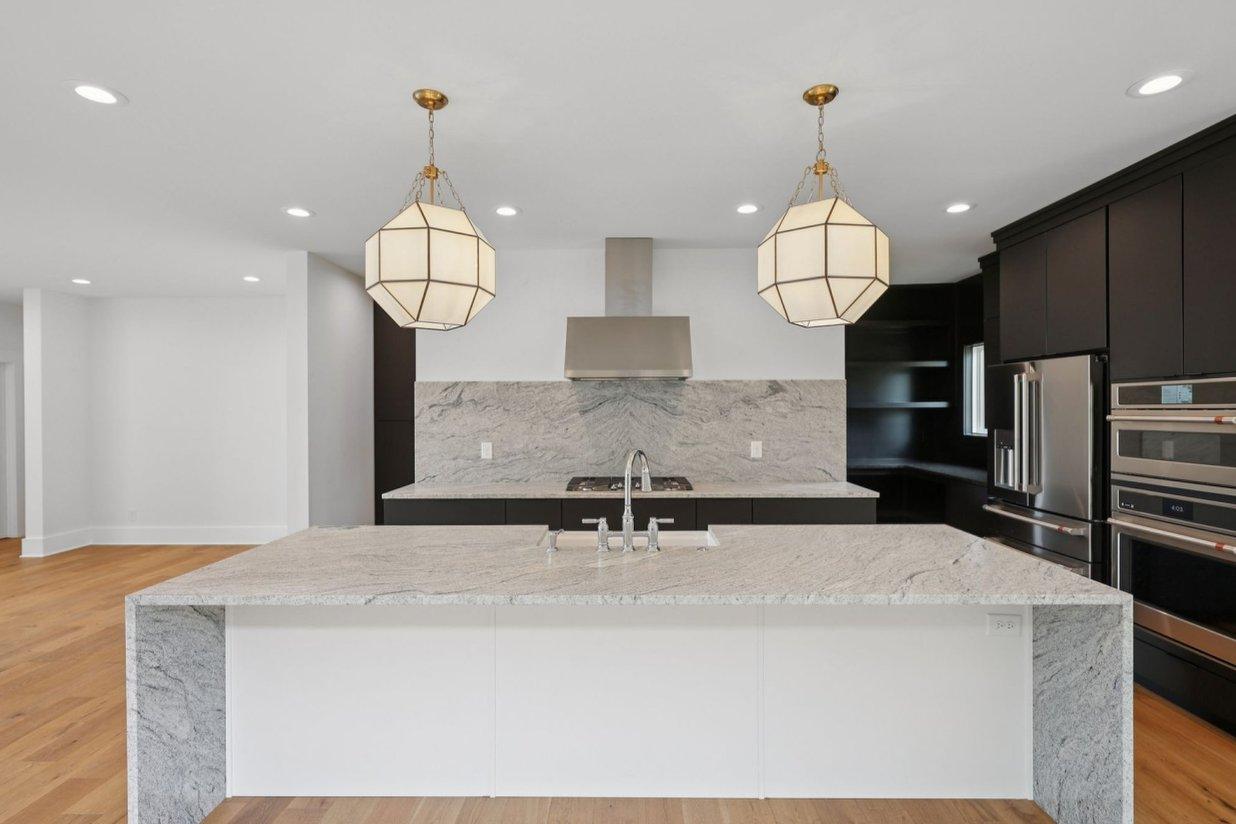 2025 photograph (before): the kitchen, a white-based granite waterfall island under two lantern pendants, black cabinets and a stainless hood.