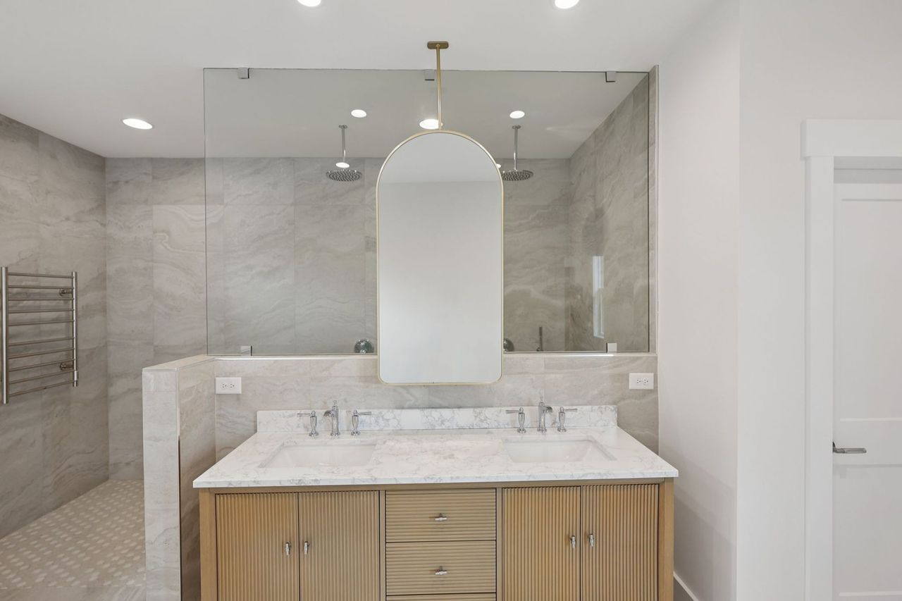 2025 photograph (before): the primary bath vanity, a fluted vanity with a white top under an arched gold mirror, gray tile behind.