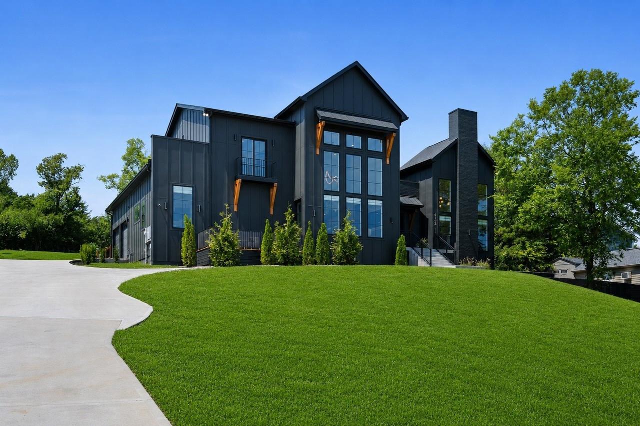 2026 photograph: Exterior Front, from driveway lawn, with black board-and-batten siding with black painted brick chimney, two-story front wall of windows with the great-room beaded chandelier visible through the glass, second-floor Juliet balcony with iron railing and orange timber brackets, front steps with black iron railings (right side)